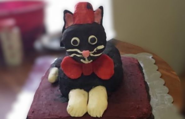 Doctor Who Fez Cat A cake shaped like a cartoonish black cat with white paws wearing a red bow tie and red fez